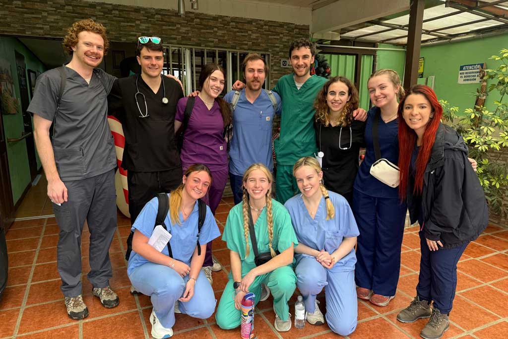 A group of ten volunteers in scrubs posing together in a clinic courtyard