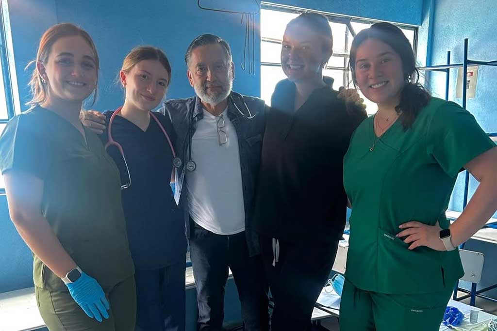Five healthcare volunteers in scrubs standing with a doctor in a clinic room