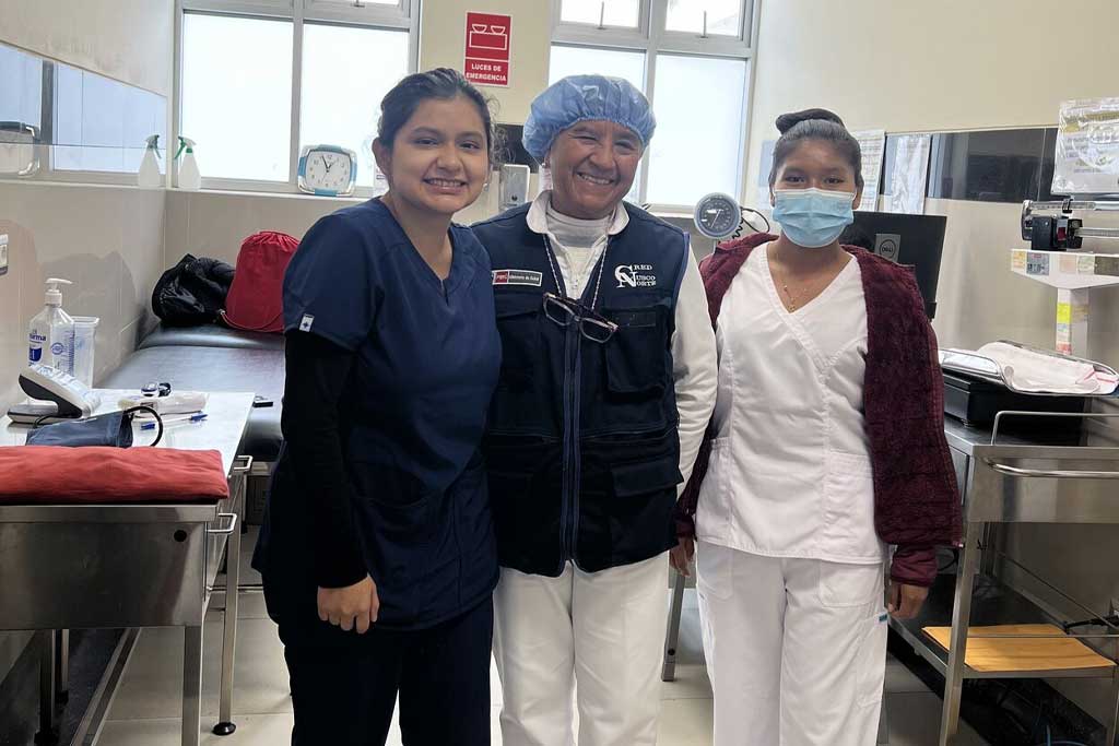 Three healthcare workers in scrubs smiling together in a clinic exam room