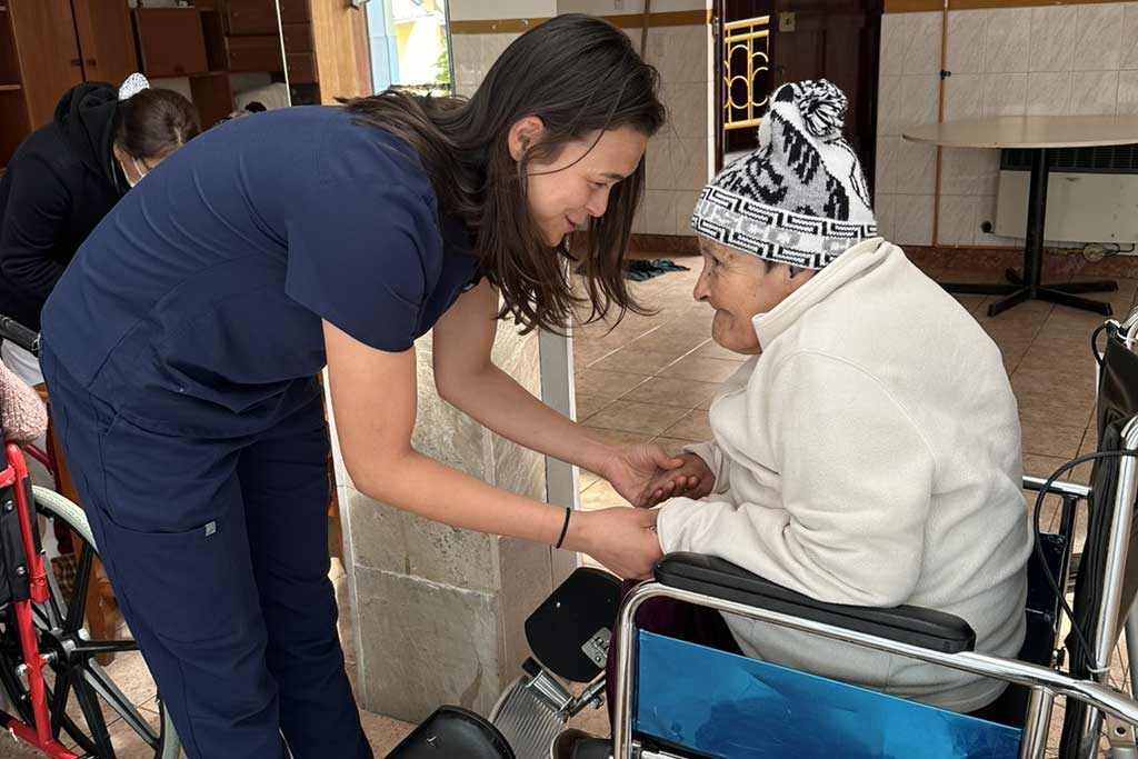 A clinician in scrubs gently holding the hands of an elderly patient seated in a wheelchair