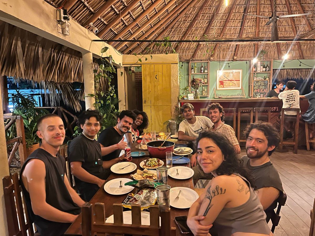 A group of students sharing dinner at a long wooden table under a palm-thatched roof