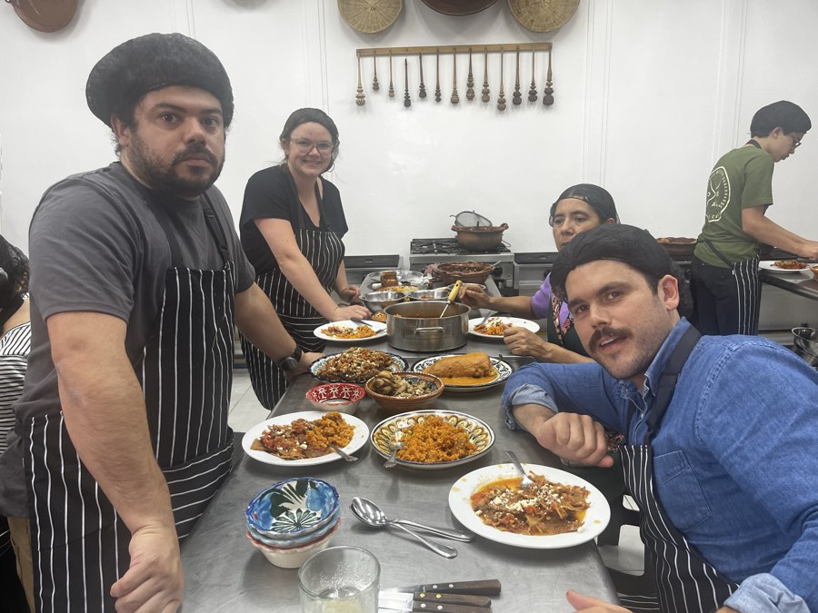 Students in aprons at a kitchen table with plates of rice, mole, and other dishes they cooked